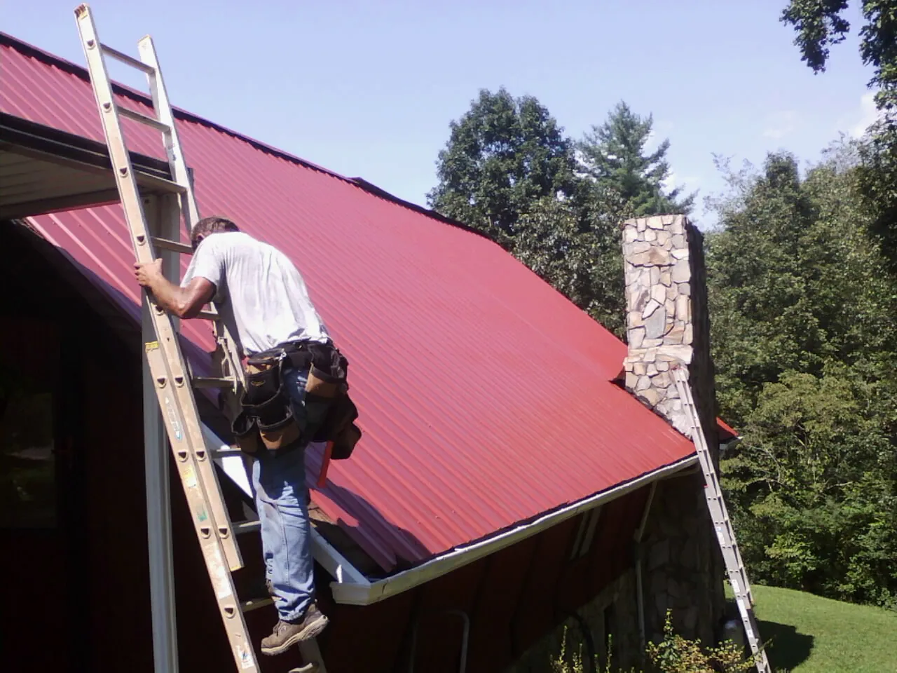 Roofer on a ladder beside a red metal roof and stone chimney — Soffit & Fascia Repair by Roofing Brothers in Township Of Washington